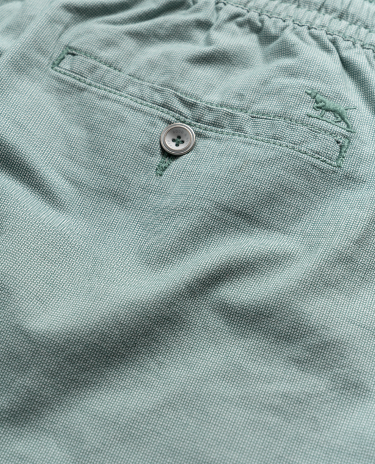 Close-up of the Rodd & Gunn Resort Short in light green fabric featuring a textured pattern. It includes a stitched buttoned pocket with an embroidered animal logo above, showcasing detail and craftsmanship. The cotton elastane blend and elastic waist offer comfort and flexibility.