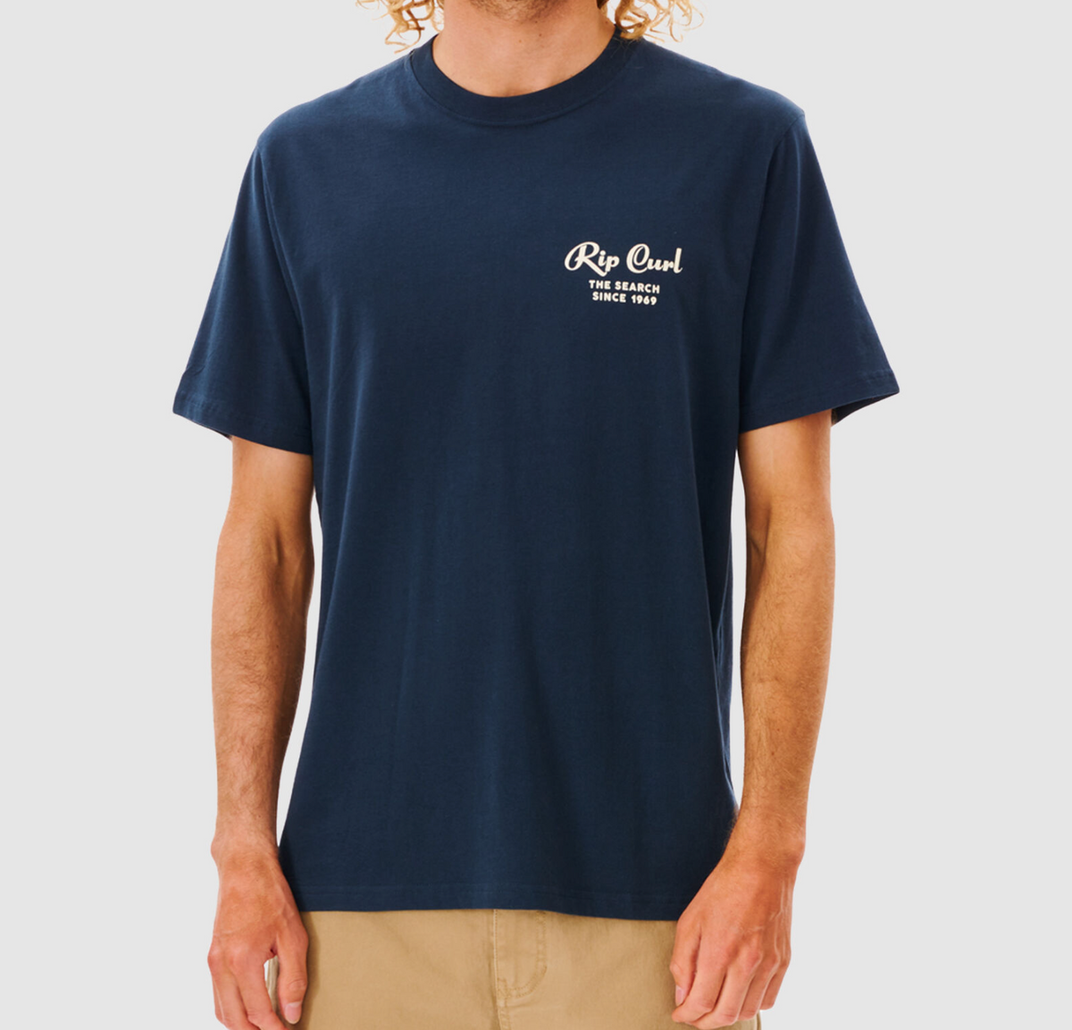 A person in a navy blue Rip Curl Twist Tee and beige shorts stands against a light gray backdrop. The comfy tee displays white text on the front: "Rip Curl The Search Since 1969.