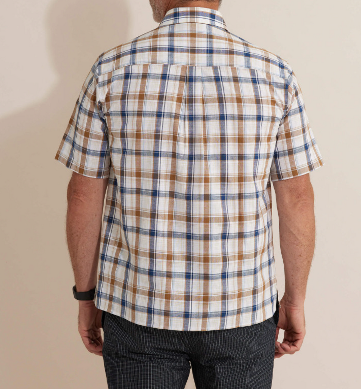 A person in a Breakaway Amber Flaxley Shirt, a comfortable short-sleeved plaid made of 100% cotton in white, brown, and blue, is seen from behind. They wear dark-textured pants and a wristwatch on their left wrist; the background is a plain light color.