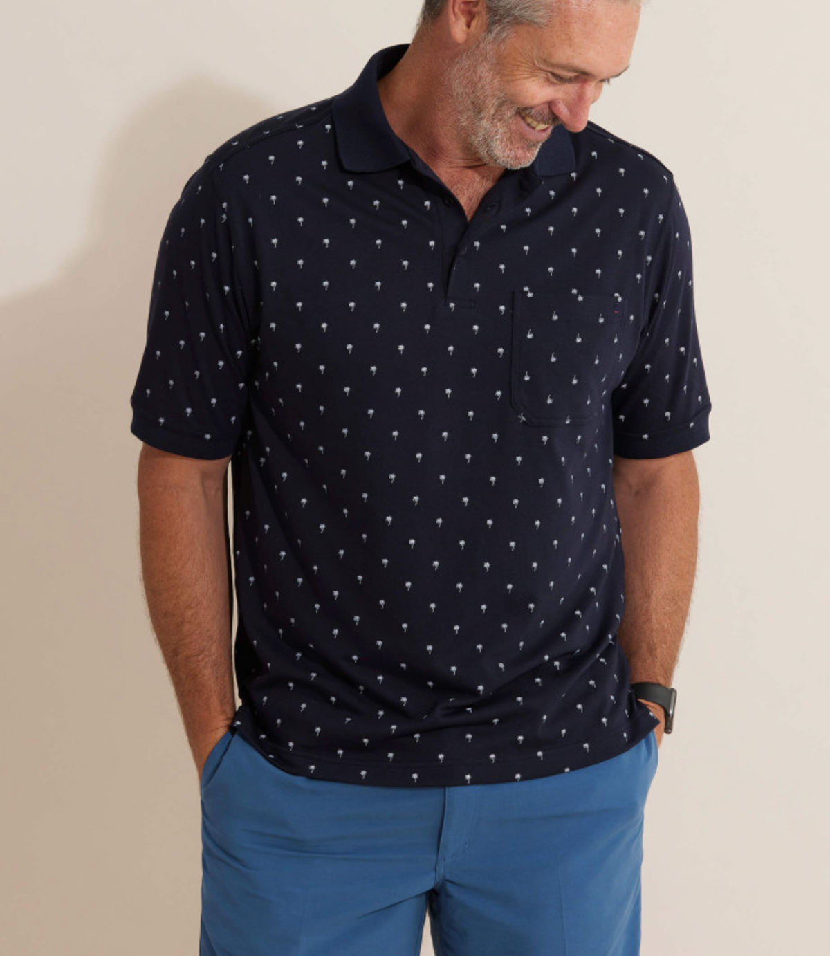 A man in a comfortable navy Breakaway Urban Polo with a subtle pattern pairs it with blue shorts. Smiling, he stands against a plain background, hands in pockets, enjoying his break from the daily routine.