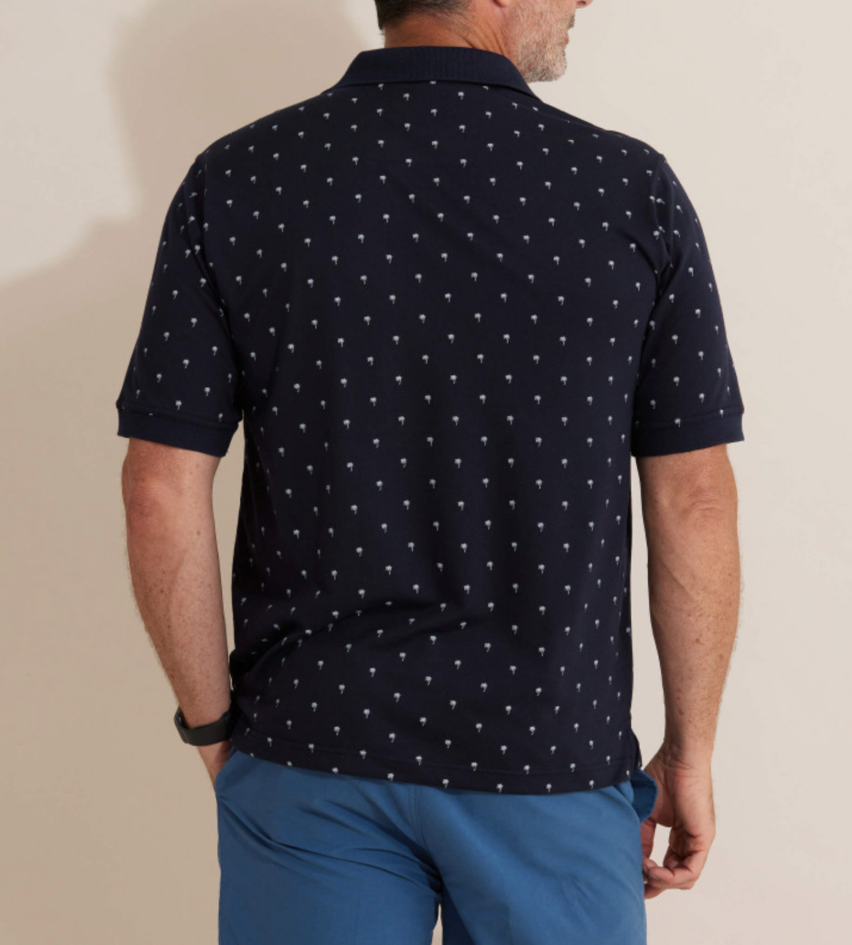 A man stands facing away, wearing the Breakaway Urban Polo in navy blue with a white palm tree pattern, rib knit collar, and paired with blue shorts. His right arm is relaxed by his side and he wears a black wristwatch.