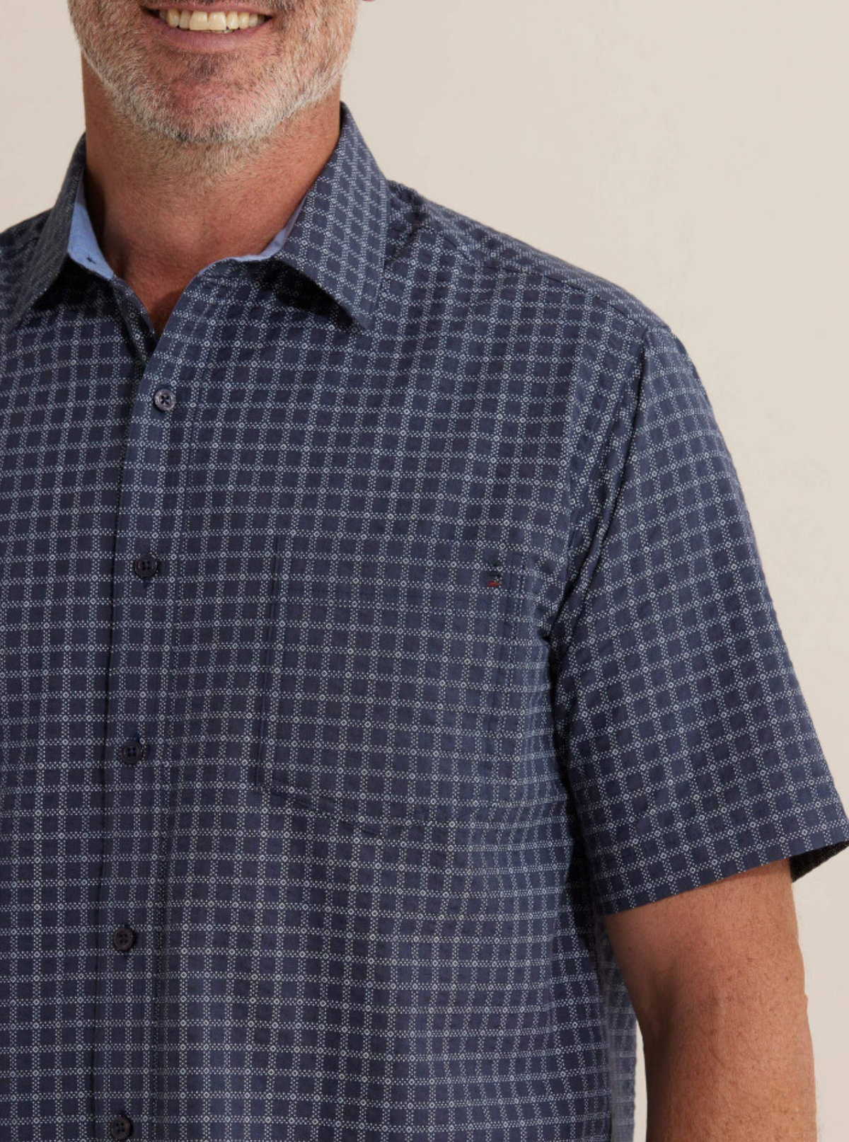 A man is wearing the Breakaway Skye Seersucker Shirt, a short-sleeved, blue patterned button-up made of 100% cotton. The photo captures his shoulders and upper arms, with a slight smile and beard against a plain background.