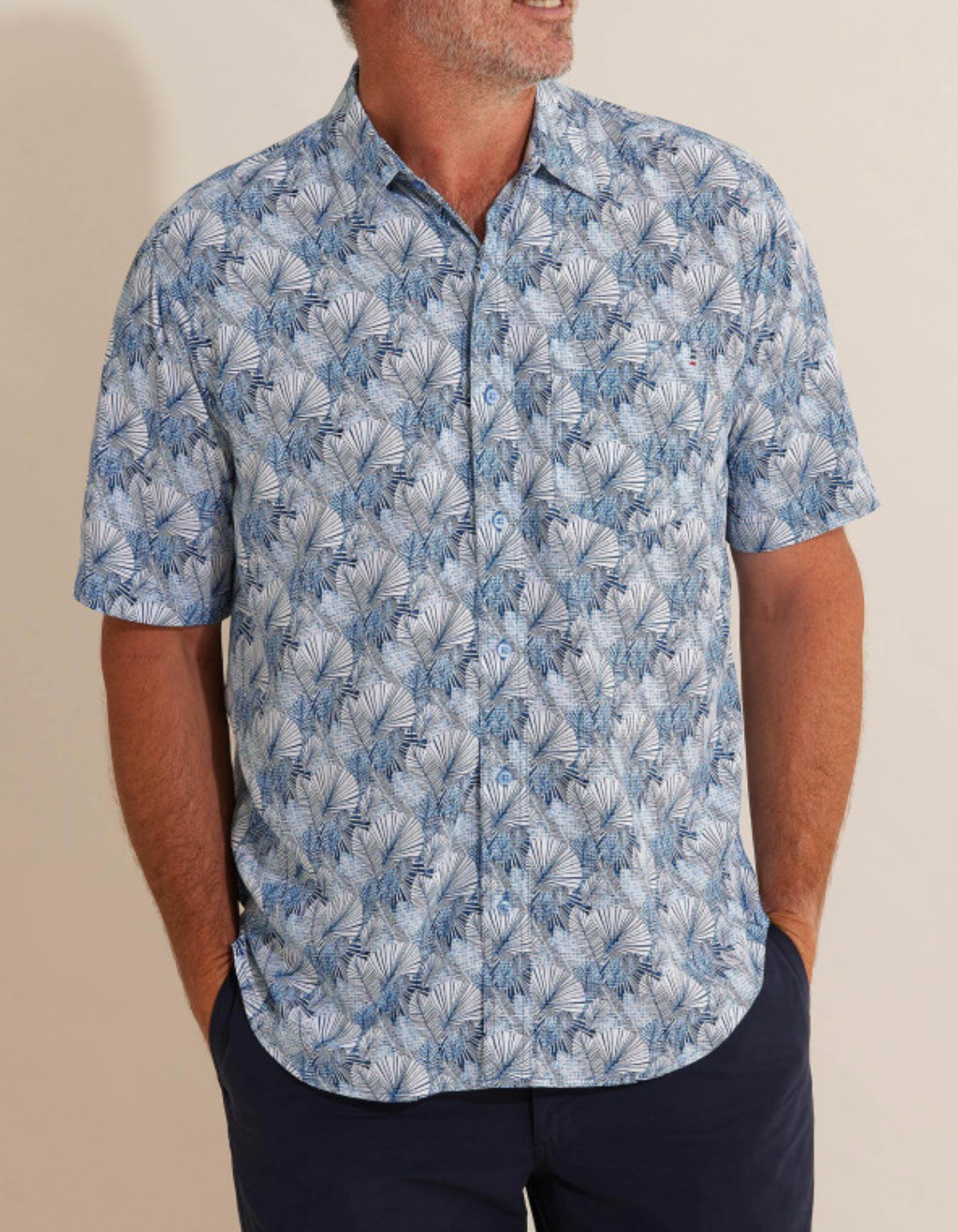 The man stands against a light background, hands in pockets, wearing a short-sleeved Breakaway Harlem Bamboo Shirt with a blue and white palm leaf pattern. Made from 100% bamboo, the shirt complements his navy blue pants perfectly.