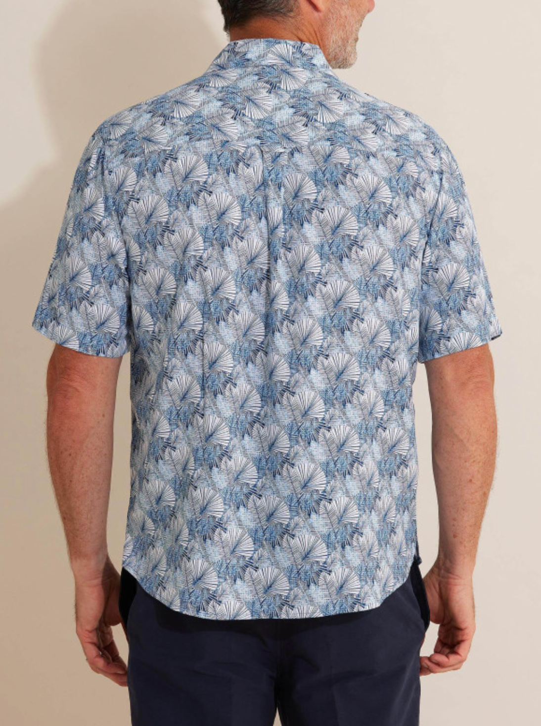 A man is pictured from behind wearing the Breakaway Harlem Bamboo Shirt, a short-sleeved, eco-friendly top featuring a 100% bamboo fabric with a blue and white abstract pattern. He pairs it stylishly with dark trousers.