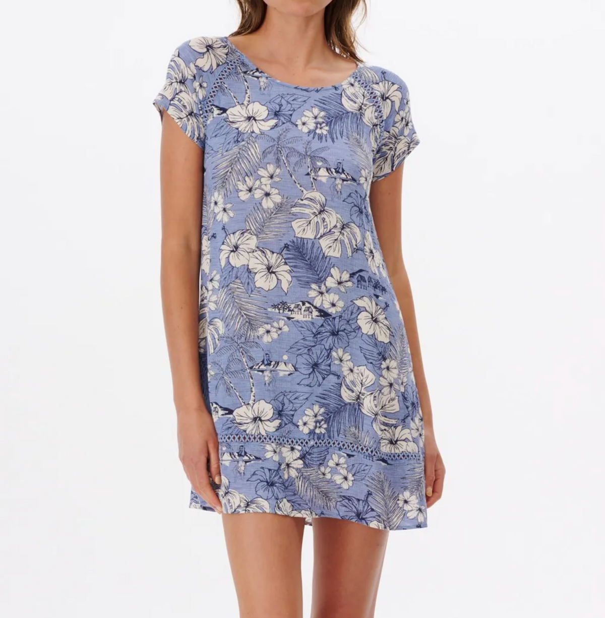 A person wears the Rip Curl Surf Treehouse Shift Dress, a blue, short-sleeved, relaxed-fit dress with a white floral pattern, subtle lace detail, and lattice insert near the hem. The background is plain white.