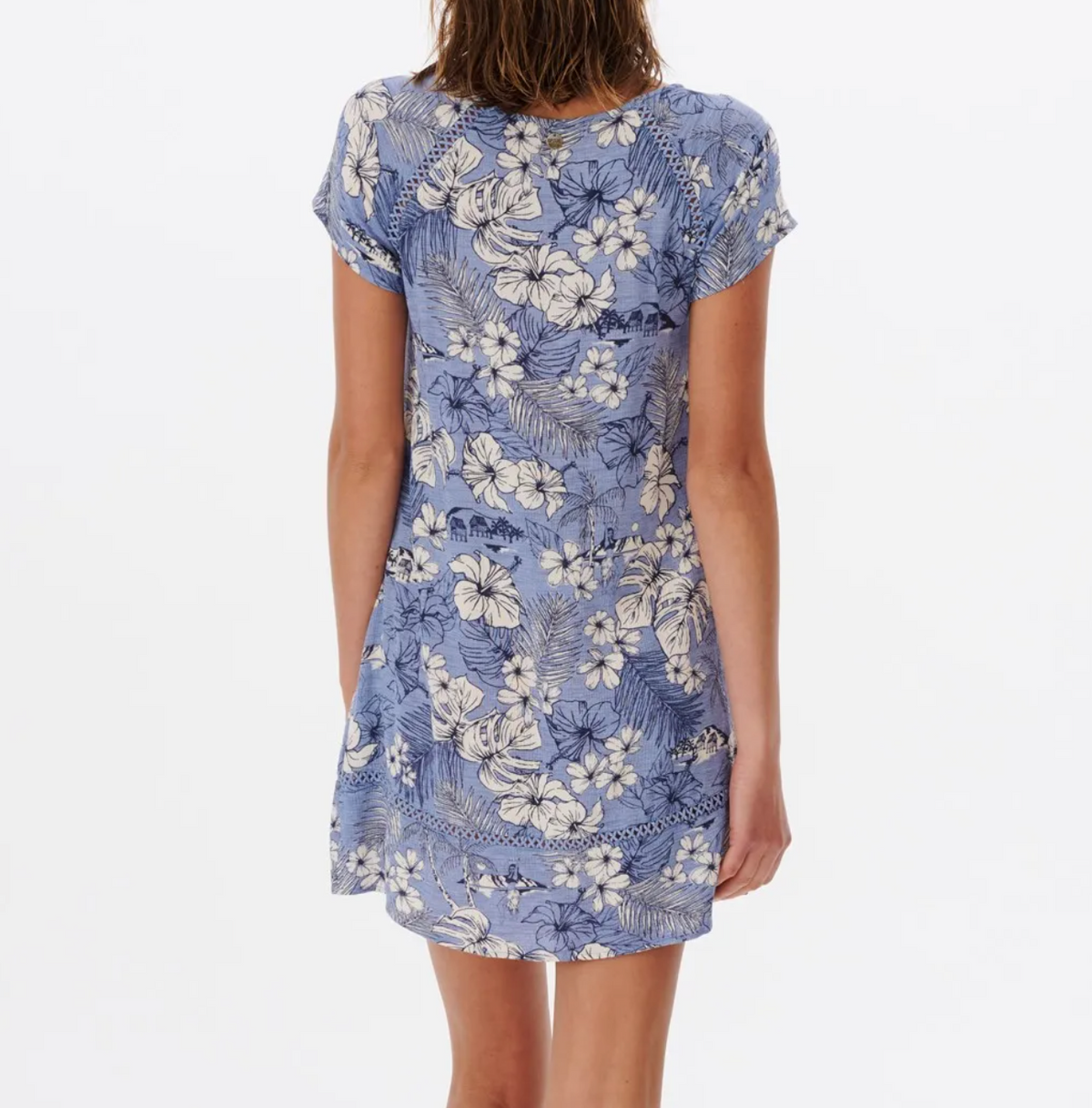 A woman in a Rip Curl Surf Treehouse Shift Dress stands with her back to the camera, highlighting a blue short-sleeved dress with a white floral pattern. Its relaxed fit flatters her frame against a crisp white background.