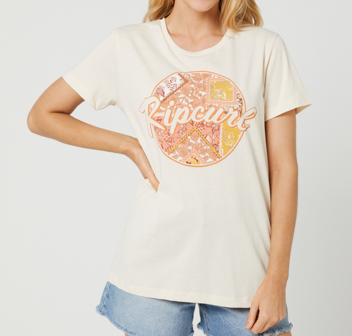 A woman models the Rip Curl Beachside Standard Tee in cream, made from 100% cotton, featuring script over a circular patterned graphic, paired with light blue denim shorts. Her left hand rests on her hip.
