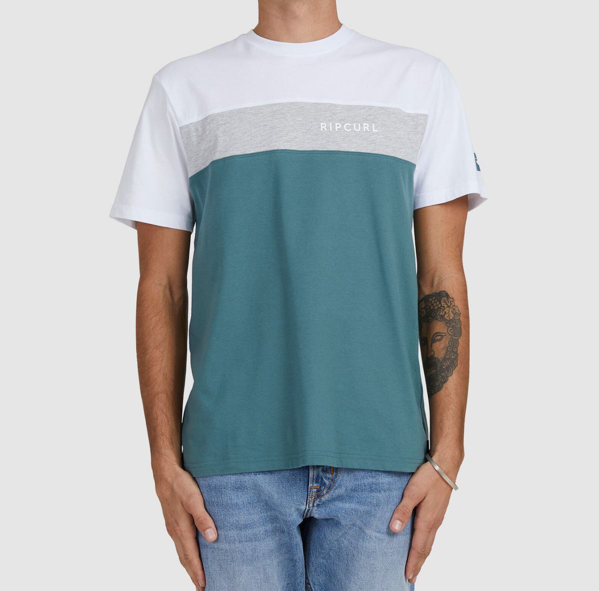 A person wears a Rip Curl Undertow Panel Tee in white, gray, and teal with "Rip Curl" on the chest. They have a forearm tattoo and pair it with blue jeans against a plain gray backdrop.