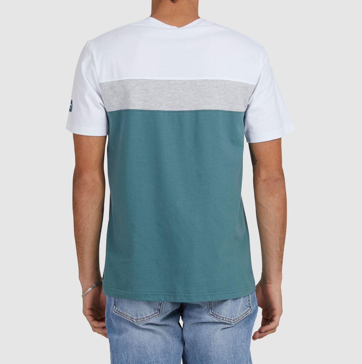 A person wearing the Rip Curl Undertow Panel Tee, featuring a short-sleeve color-block design in white, gray, and teal, stands with their back to the camera. It's paired with light blue jeans against a plain white background for a casual and comfortable look.