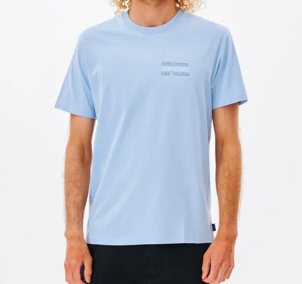 The individual wears a light blue Rip Curl Revival Repeater Tee made from 100% Cotton Jersey with subtle text on the left chest. They're paired with black pants and stand against a white background, arms at their sides, complementing their curly blond hair.
