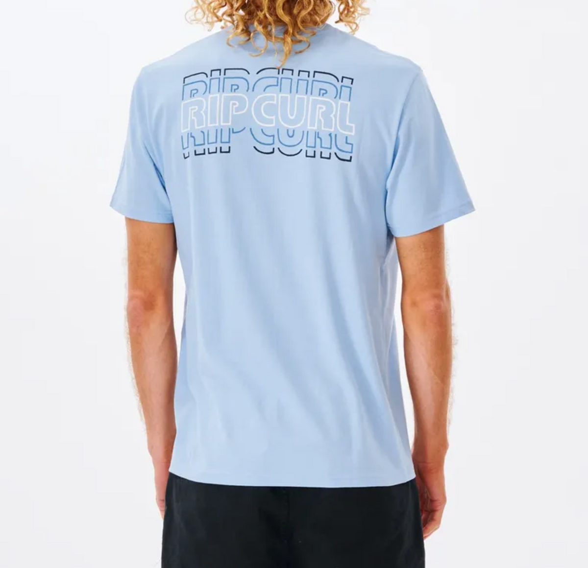 Wearing the light blue Rip Curl Revival Repeater Tee, made from 100% Cotton Jersey, with its iconic logo stylishly layered on the back, a person with curly hair pairs it with dark shorts against a plain white background, showcasing effortless style and comfort.