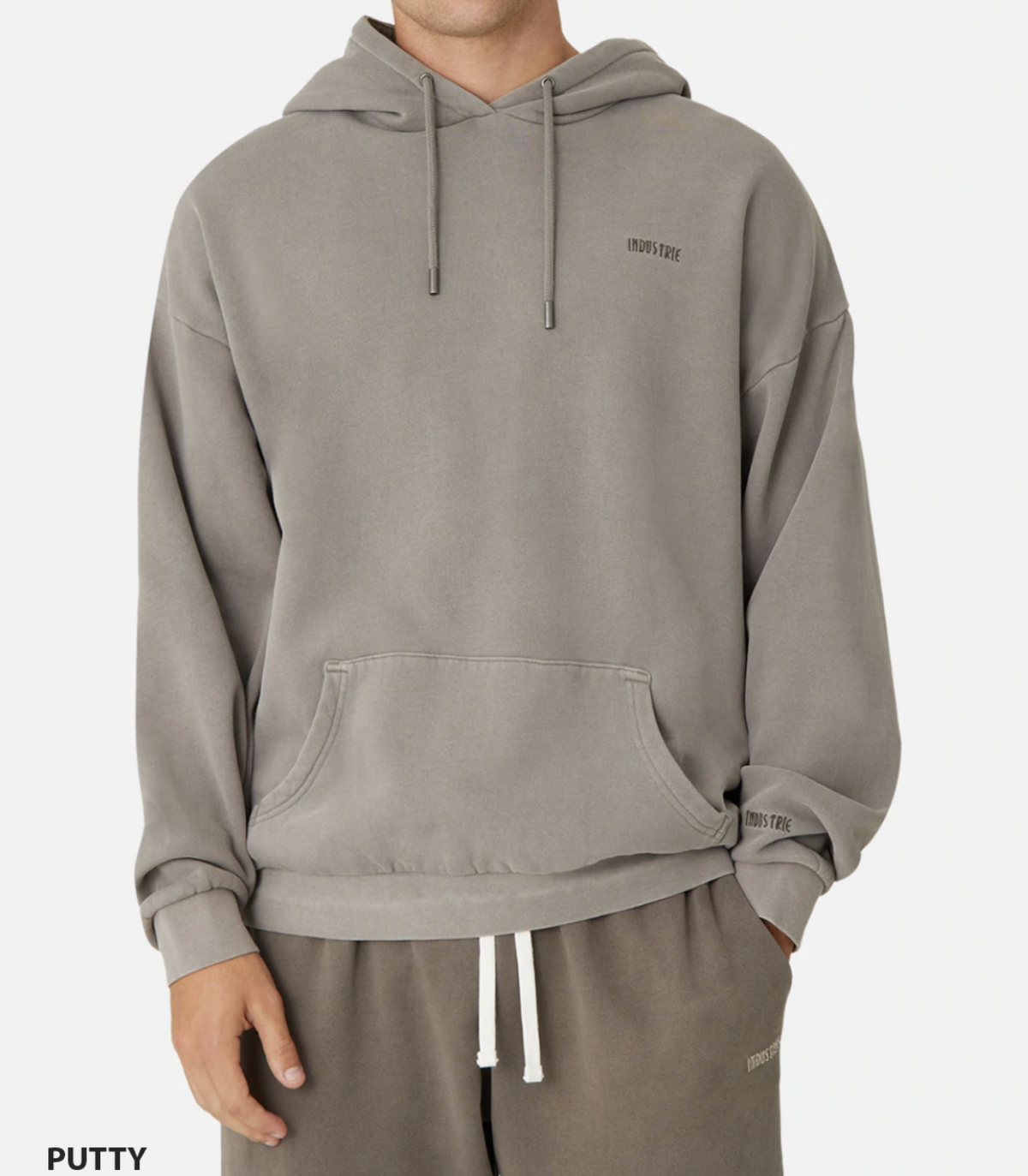 A person is wearing the Industrie The Del Sur Hoodie in gray with an embroidered chest logo and matching sweatpants. This comfy oversized hoodie has a drawstring hood and front pocket. The crisp white backdrop enhances its casual appeal.
