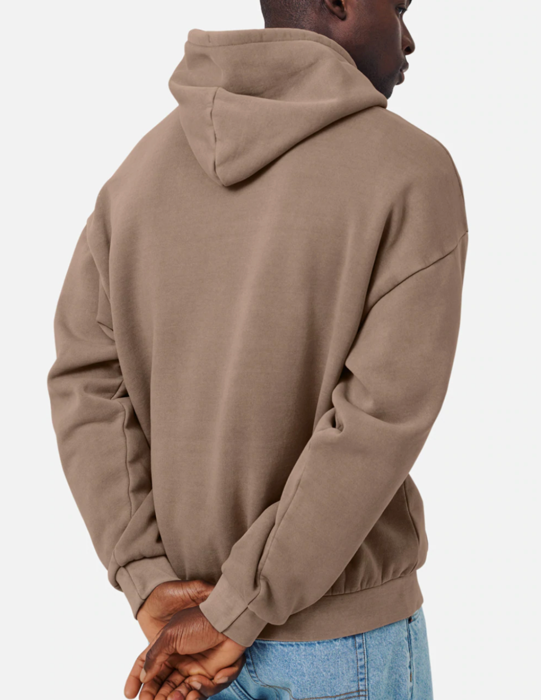 A person wearing an oversized Industrie The Del Sur Hoodie in tan and light blue jeans faces away, arms folded behind them, with the hood up against a plain white background.