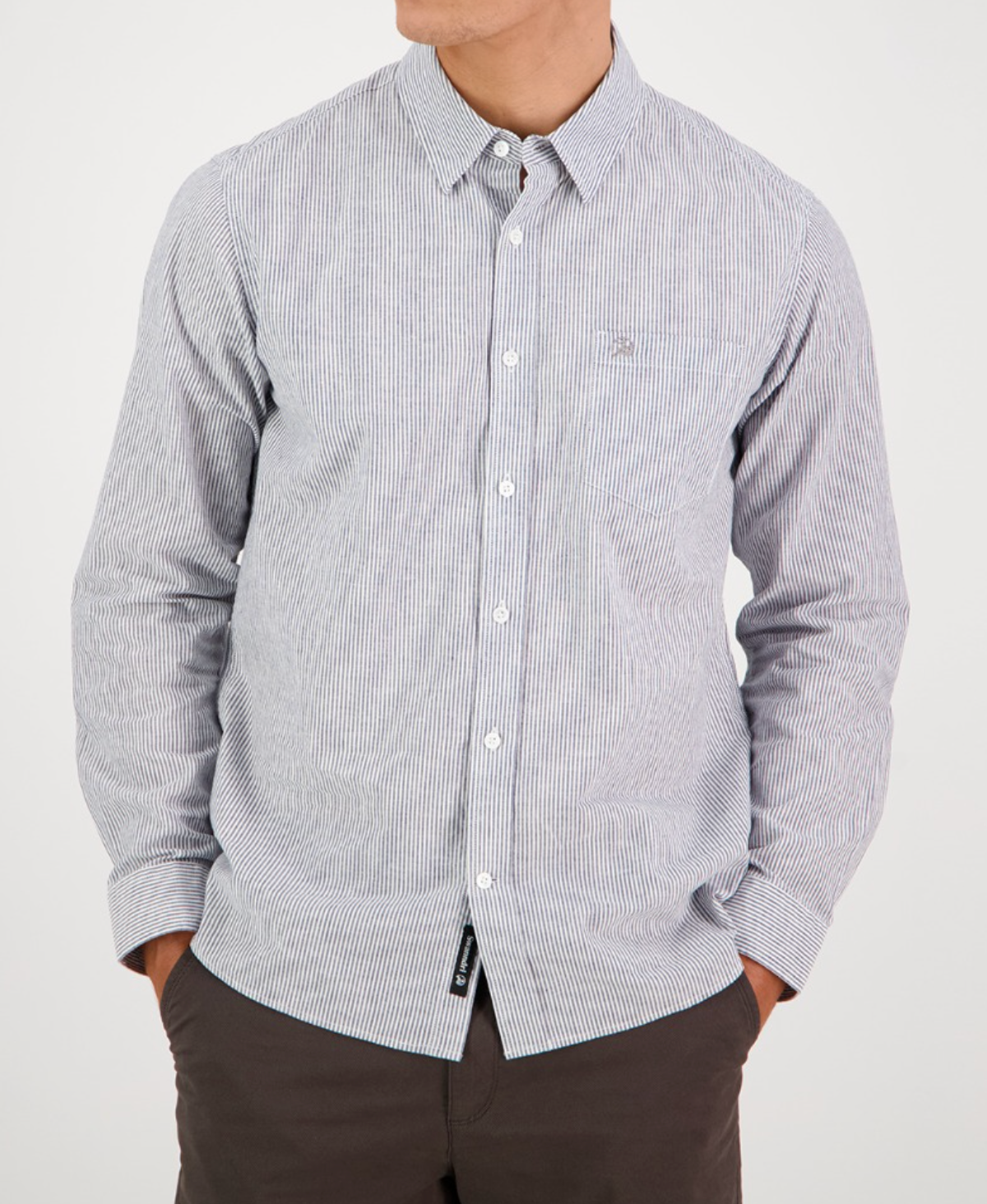 A person models the Swanndri Carillo Beach L/S Shirt, featuring light blue and white stripes in a tailored fit with long sleeves. They pair it with dark pants, hands in pockets for a comfortable vibe, against a plain background while their face is out of view.