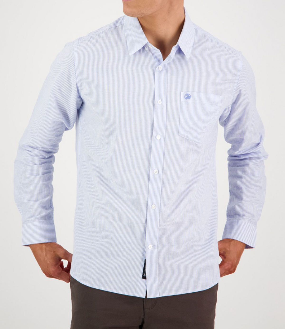 A person in a tailored fit Swanndri Carillo Beach L/S Shirt, light blue with stripes and a left pocket, stands against a white background. Their hands rest on their hips, highlighting the shirt's comfortable, casual style.