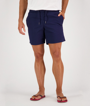 A person wearing a white shirt, Swanndri Somerset Beach Short in navy blue cotton, and red and black flip-flops. They stand against a plain white background, hands tucked into the pockets of the comfortable shorts.