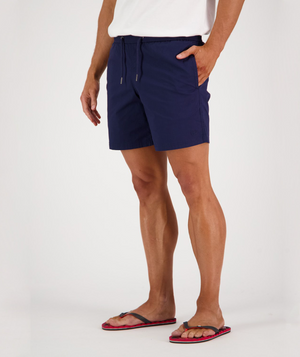 A person wearing a white t-shirt, Swanndri Somerset Beach Short with drawstrings, and flip-flops stands with one hand in their pocket against a plain white background.