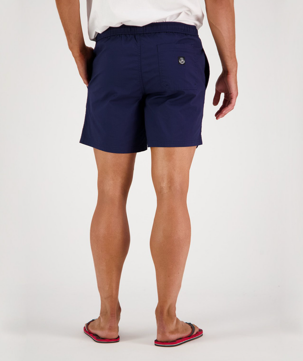 View from behind: a person dressed in a white shirt, Swanndri Somerset Beach Short in dark blue, and flip-flops. The shorts are designed for ease and style with an emblem on the back pocket. The individual stands against a plain white background.