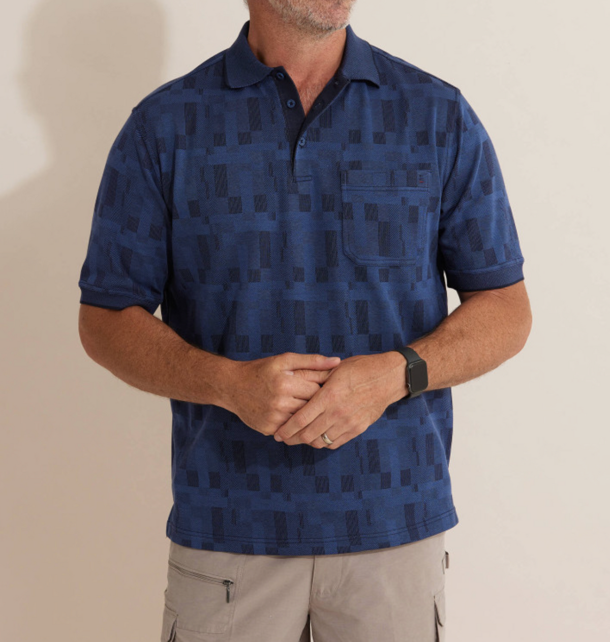 A man stands against a plain light background wearing the Breakaway Jacquard Micro Prima Polo, a comfortable blue patterned shirt paired with beige cargo shorts. A smartwatch adorns his wrist as he casually clasps his hands in front.