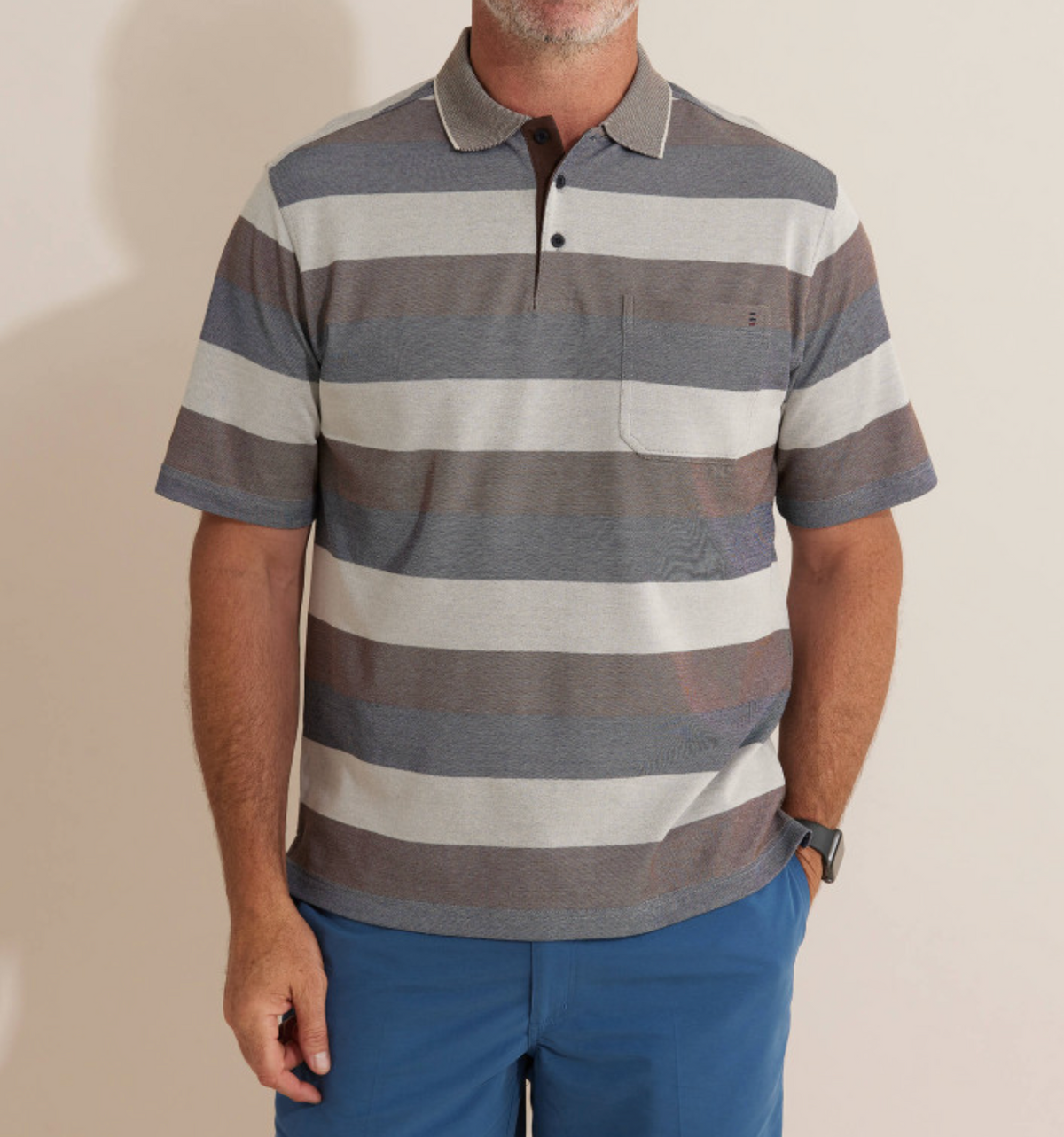 A man wearing the Breakaway Machu Micro Prima Polo with gray and brown horizontal stripes, a rib knit collar, chest pocket, and blue pants stands against a light background.