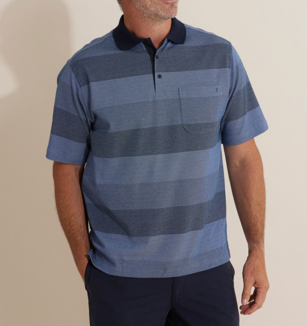 A man in the comfortable Breakaway Inva Micro Prima Polo, featuring blue horizontal textured stripes and a navy collar, pairs it with dark pants. With short hair and a beard, he casually places his left hand in his pocket while looking to the side.