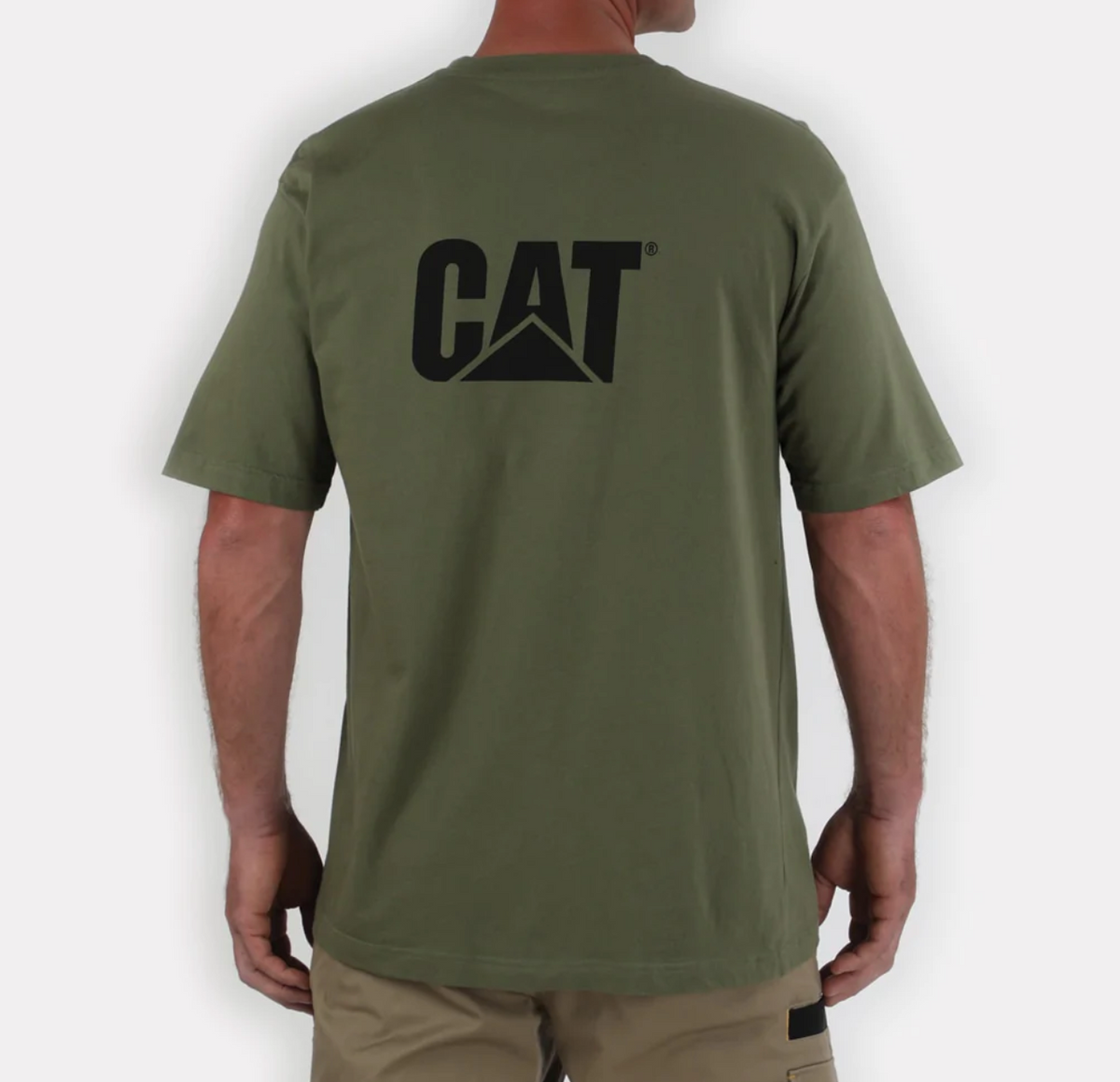 A person stands with their back to the camera wearing a CAT Trademark Tee, made of 100% cotton in green, featuring bold black "CAT" lettering on the back. They pair it perfectly with beige shorts against a plain white background.