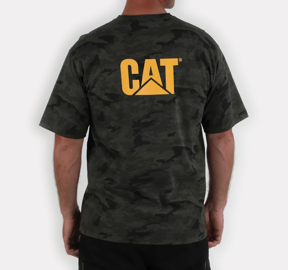 A person is wearing the CAT Trademark Tee, a dark camouflage T-shirt made from 100% cotton, with the iconic yellow "CAT" logo on the back. The casual tee contrasts with a plain, light-colored background.