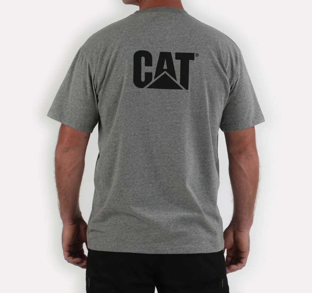 Wearing the casual CAT Trademark Tee, featuring a bold black CAT print on the back, a person stands in dark pants against a plain white background. This 100% cotton gray shirt is viewed from behind.
