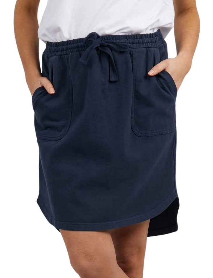 The person is wearing the Foxwood Palm Skirt in navy blue, which features a knee-length design with front pockets and an elastic waistband tied with a drawstring. The skirt's hemline is slightly curved, and it is made from 100% Cotton Baby Cord for extra comfort. They are also wearing a white top and have their hands in the skirt's pockets.
