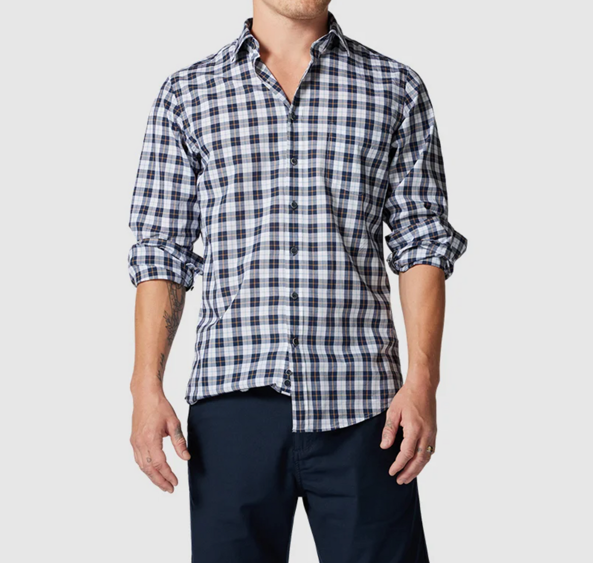 Wearing the Rodd & Gunn Mount Albert Sports Fit Shirt, a stylish slim-fit blue and white checkered 100% cotton piece, person stands against a plain background. The shirt features rolled-up sleeves, is buttoned up, and they pair it with dark pants, hands relaxed by their sides.
