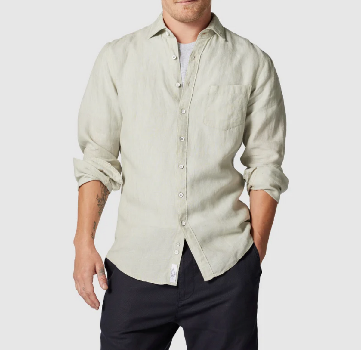 A person in a light beige Rodd & Gunn Coromandel Sports Fit Shirt, 100% linen with rolled-up sleeves and chest pocket, stands against a plain background. They wear dark pants, have a forearm tattoo, and a white t-shirt underneath.