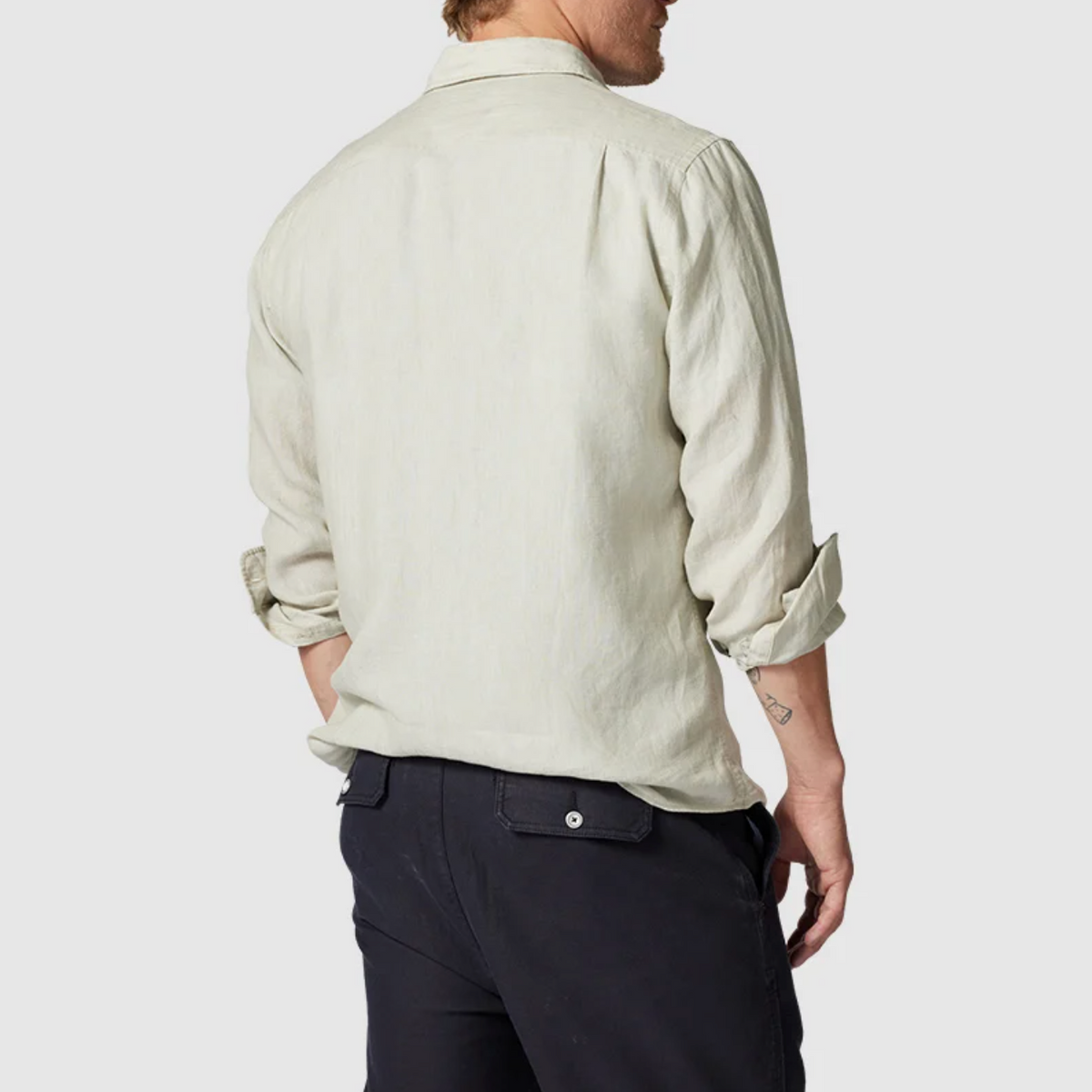A person with short hair and a small arm tattoo is seen from behind wearing a Rodd & Gunn Coromandel Sports Fit Shirt made of 100% linen, featuring rolled-up sleeves and paired with dark pants against a light gray background.