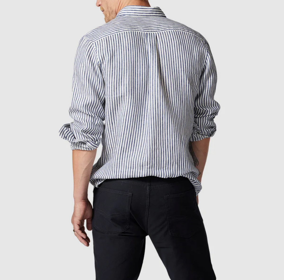 A person wearing a Rodd & Gunn Port Charles Sports Fit Shirt in blue and white stripes with black pants, viewed from the back. The long sleeves of this 100% linen shirt are slightly rolled up. The background is plain light gray.