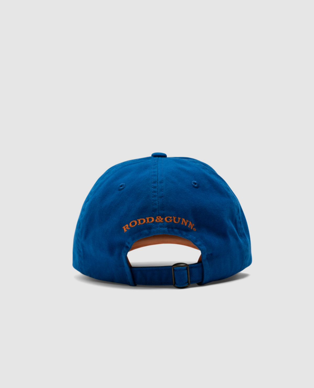 A blue Rodd & Gunn Gunn Cap is shown from the back, highlighting the "RODD & GUNN" embroidered logo in bold orange. Made from durable cotton twill, this cap includes an adjustable strap with a metal buckle on a light gray background.