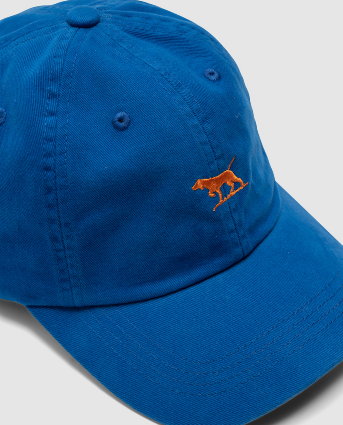 The Rodd & Gunn Gunn Cap is a blue cap with an embroidered orange dog on the front, crafted from heavy-duty cotton twill. It features seam detailing, a curved brim, and the classic Rodd & Gunn Embroidered Logo against a plain white background.
