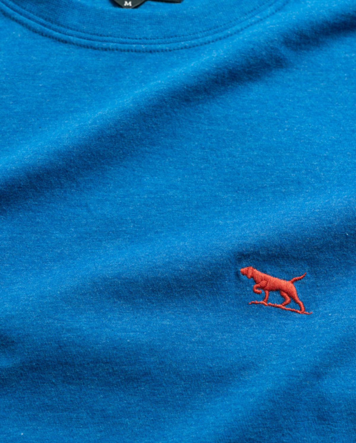 The Rodd & Gunn "The Gunn T-Shirt" in blue, made from premium Australian Cotton, features a small red dog logo and a sports fit for comfort. The medium size is indicated by the "M" visible on the tag.