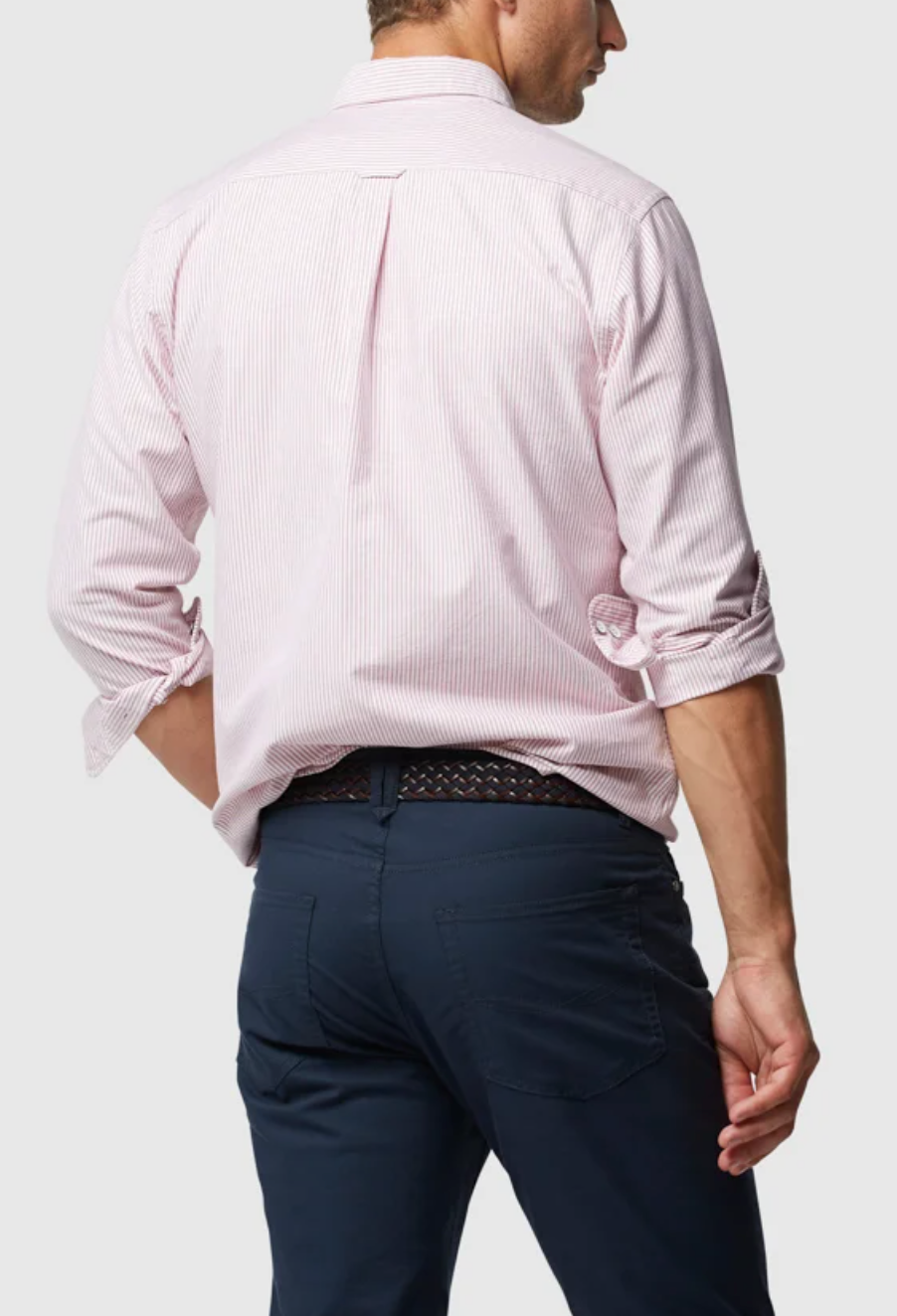 With their back to the camera, a person wears a light pink Rodd & Gunn Oxford Stripe Sports Fit Shirt made from 100% cotton. The sleeves are rolled up, paired with dark blue pants, and their left hand rests in the pocket, highlighting a stylish patterned belt.