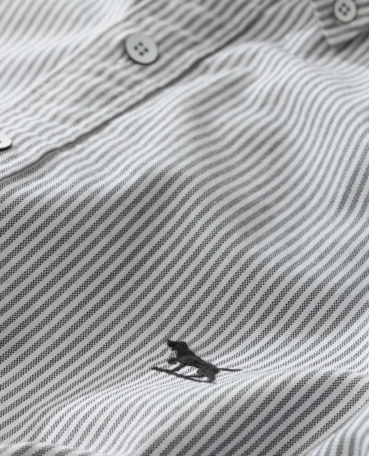 Close-up of a Rodd & Gunn Oxford Stripe Sports Fit Shirt, featuring its gray and white striped 100% cotton fabric. Small buttons enhance the sports fit, while an embroidered animal logo near the hem adds charm. The texture and evenly spaced stripes showcase its quality craftsmanship.