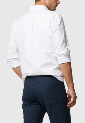 A man in a white Rodd & Gunn Gunn Oxford Sports Fit Shirt, made from 100% cotton, stands with his back turned. The sports fit design has rolled-up sleeves that complement his navy pants against the plain light gray background.