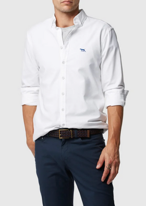 A person wears a Rodd & Gunn Gunn Oxford Sports Fit Shirt in 100% cotton with rolled-up sleeves, navy pants, and a small blue embroidered chest logo. They accessorize with a patterned belt against a plain light gray background.