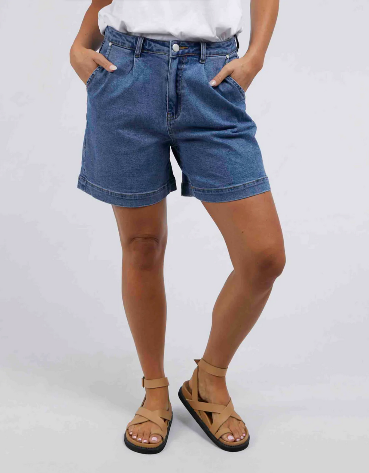 A person wearing a white shirt, Foxwood Melody Shorts made of soft stretch denim, and tan sandals stands with hands in their pockets against a plain background.