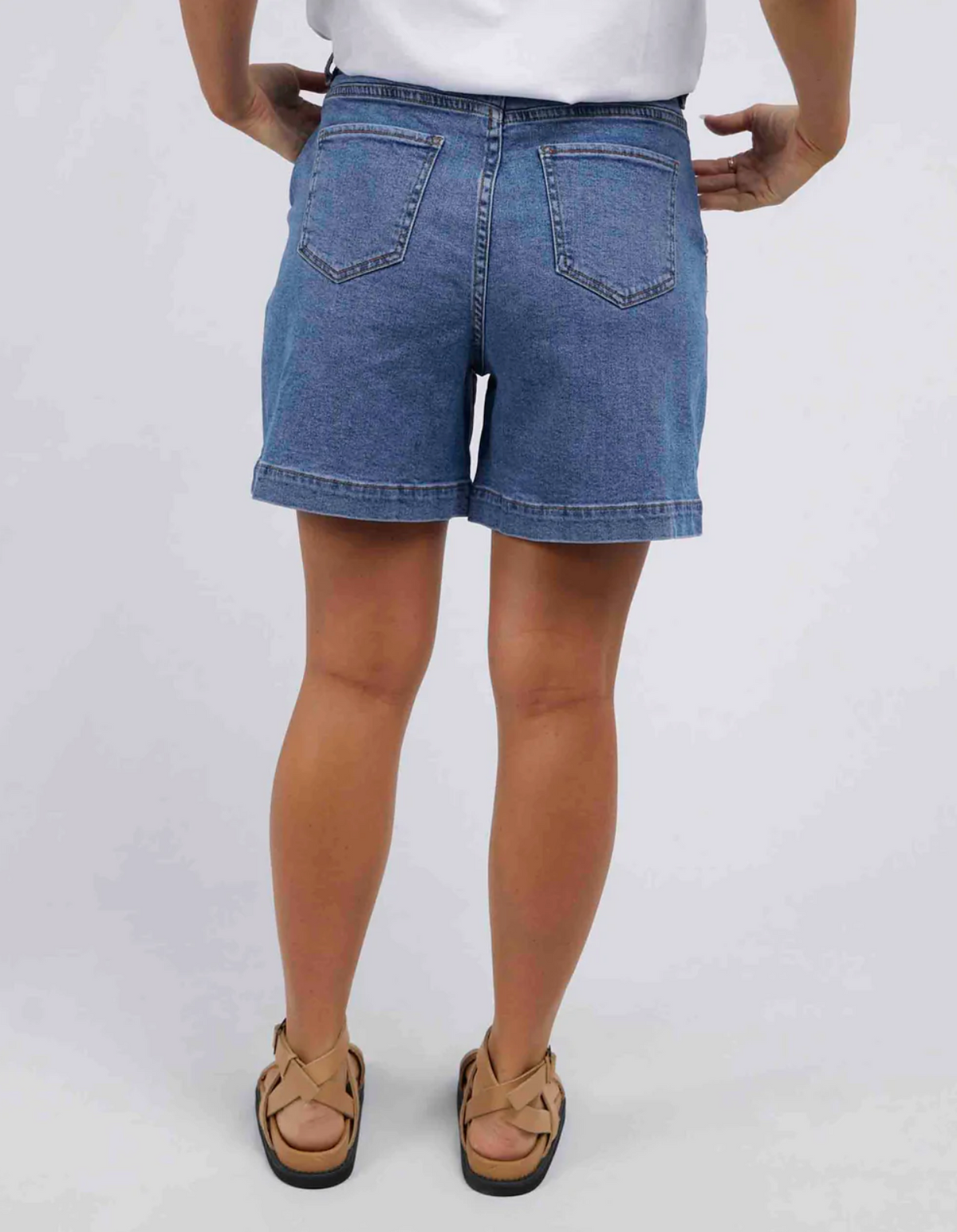 Rear view of a person in Foxwood Melody Short, a white shirt, and matching tan sandals. The soft stretch denim with back pockets ensures comfort as they stand with hands on hips, set against a plain white background.