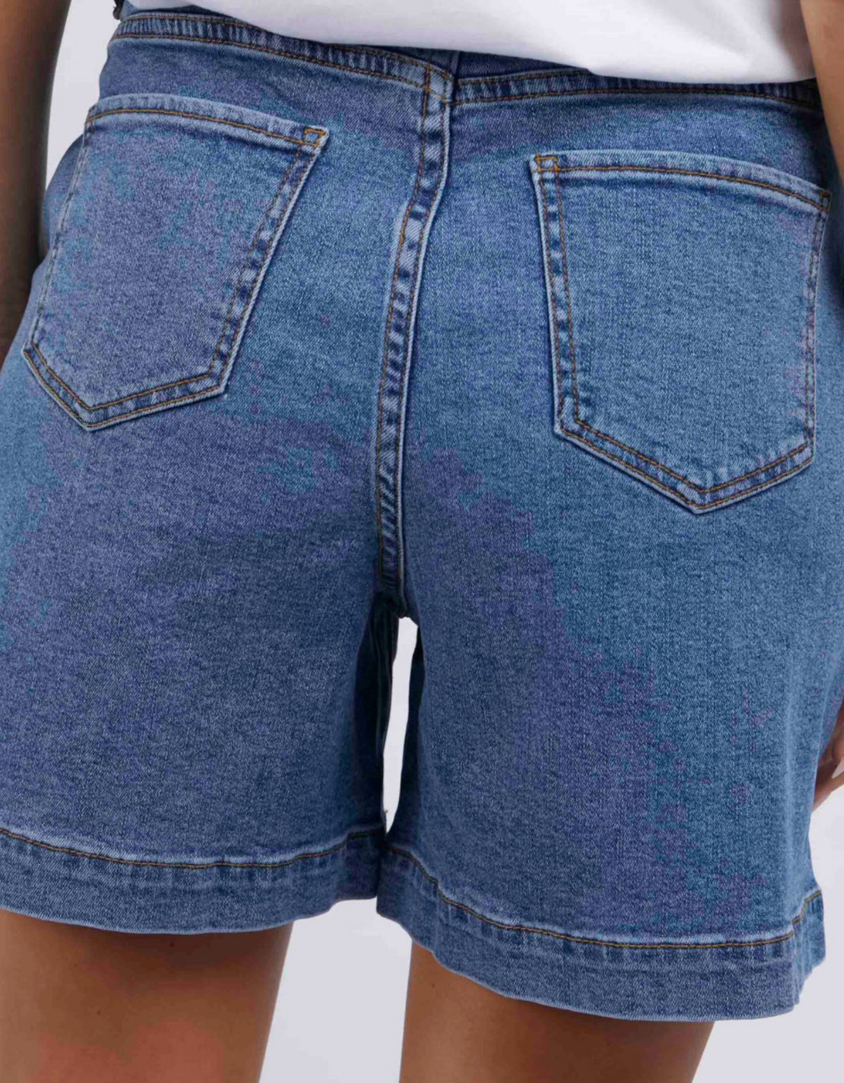 A person is wearing Foxwood Melody Short, crafted from soft stretch denim in blue with back pockets, paired with a white top. The image emphasizes the back view of these standard-fit shorts, highlighting their design and stitching.