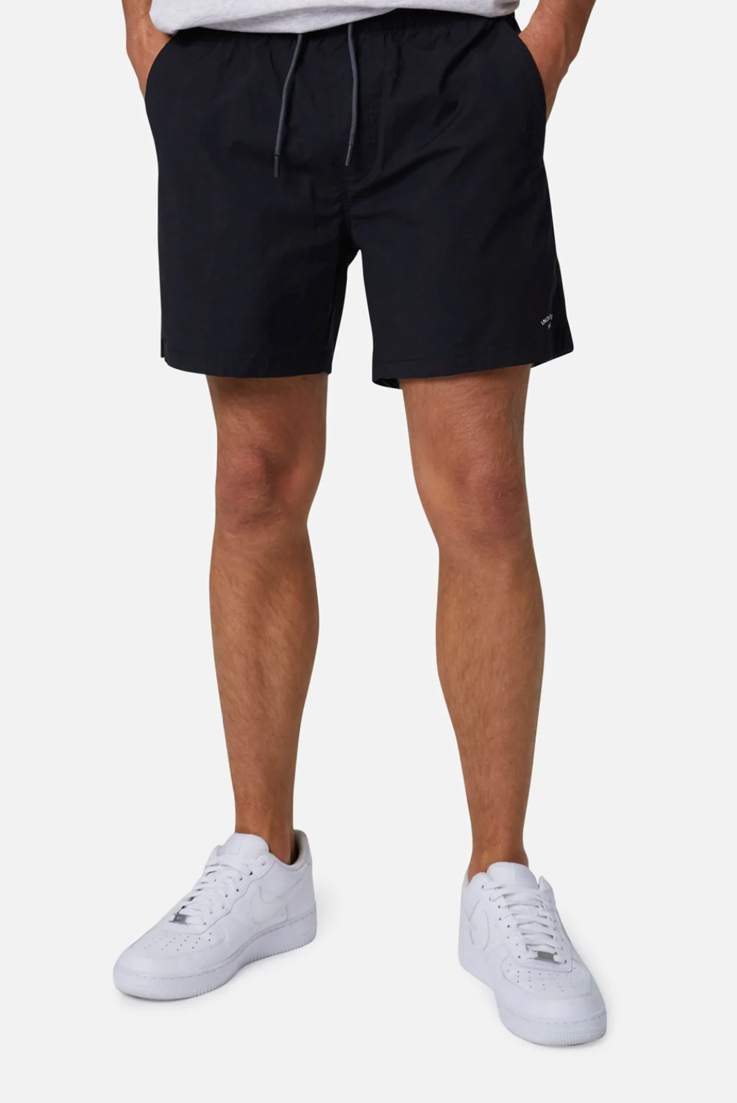 A person wearing Industrie The Bahama Angler Short in black, featuring an elastic waist and hands resting in the pockets. They are also sporting white sneakers and a light-colored top, with the lower part of the top visible. The background is plain white.