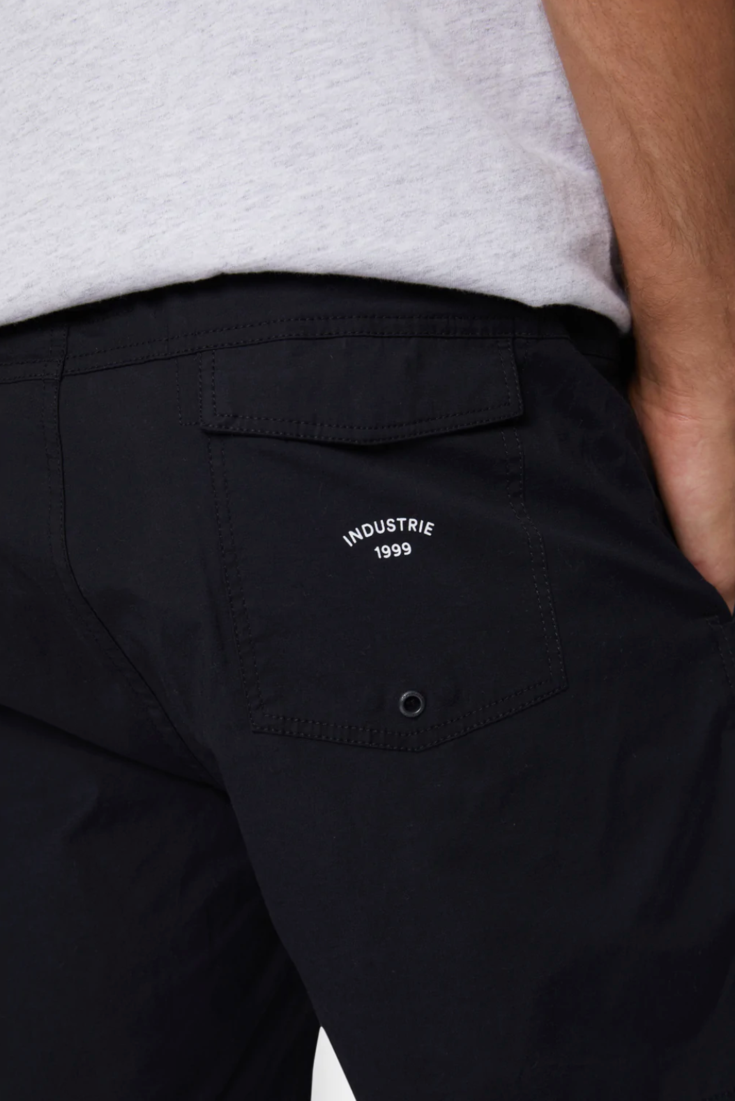 Close-up image of a person wearing Industrie The Bahama Angler Short in black, featuring a back pocket with the Industrie logo and "1999" printed above it. The person is also wearing a light-colored shirt, with their hand partially visible in the frame.