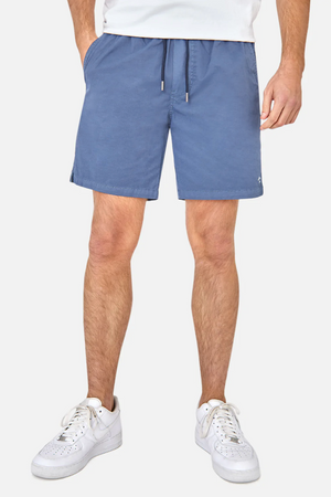 A person stands against a white background wearing a white T-shirt, white sneakers, and Industrie The Bahama Angler Short—blue swim shorts featuring an elastic waist. The focus is on the Industrie logo on the shorts and their legs.