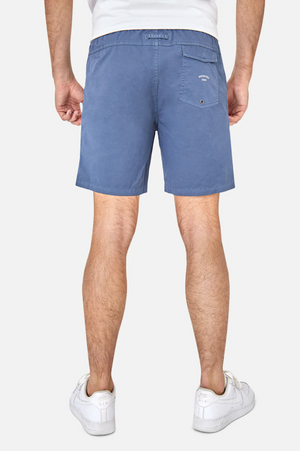 Wearing pastel blue Industrie The Bahama Angler Shorts with an elastic waist and white sneakers, a person stands against a plain white background. The Industrie logo on the shorts' back pocket complements a glimpse of their white t-shirt.