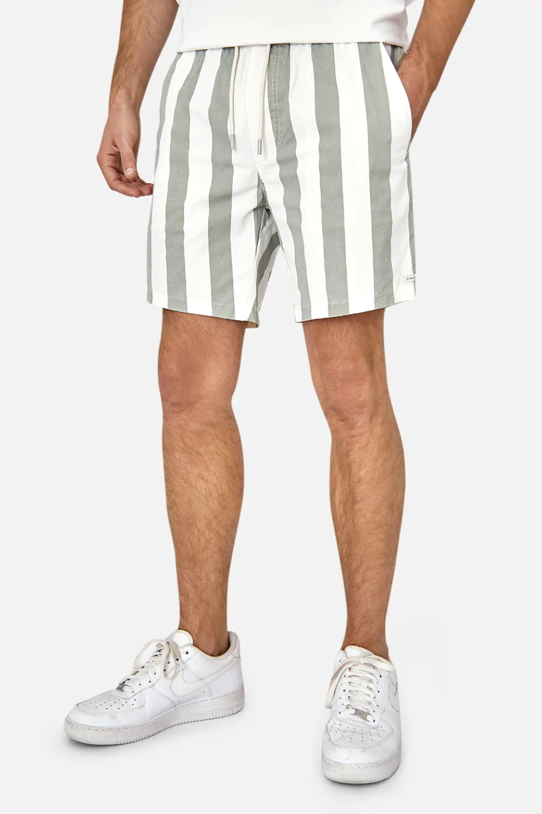 An individual dons Industrie The Beverly Bahama Short, featuring green and white vertical stripes with a drawstring and elastic waist. They wear white sneakers; the upper body is mostly cropped out, set against a plain light gray backdrop.