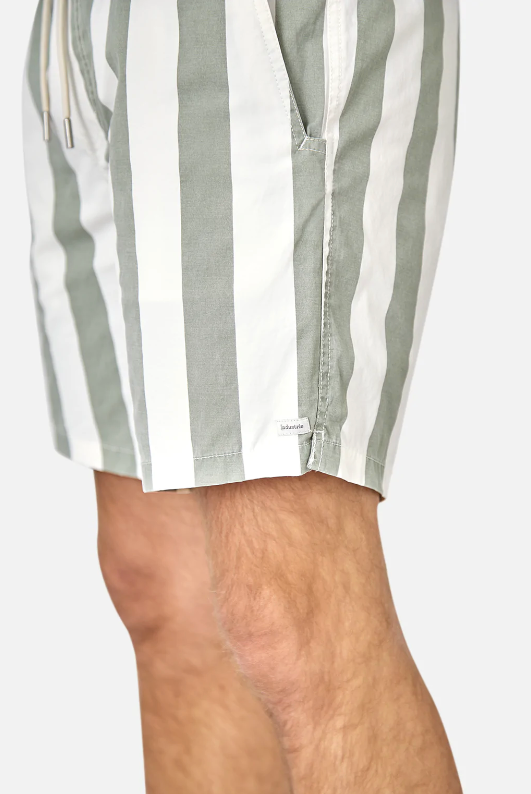 A close-up shows a person wearing Industrie The Beverly Bahama Short in pastel green and white vertical stripes, featuring side pockets and a drawstring elastic waist. The subtle Industrie logo stands out against the plain, light gray background.