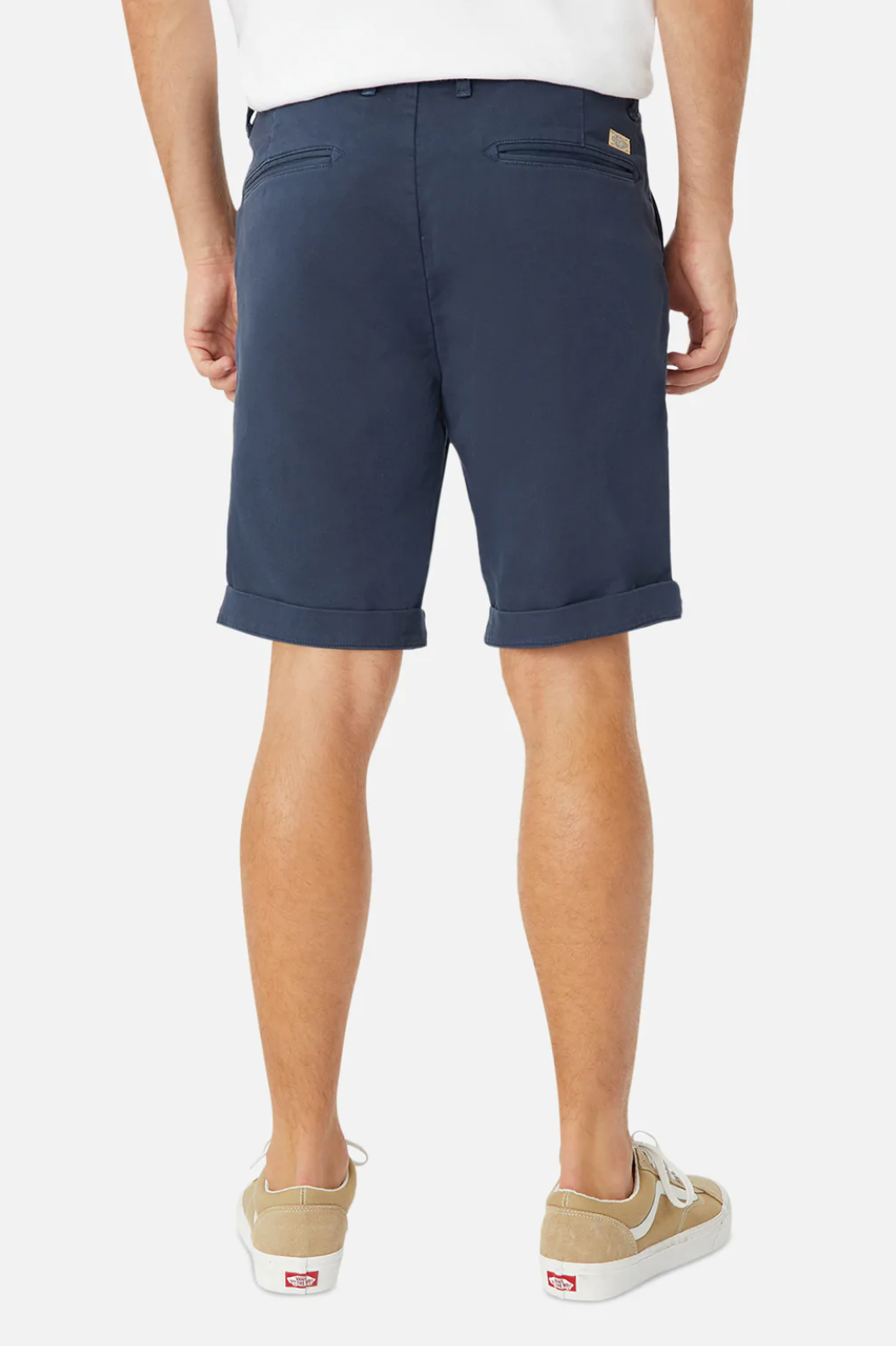 A person stands facing away, donning a relaxed fit white shirt with navy blue Industrie The Rinse Drifter Shorts made of cotton elastane, featuring rolled cuffs, paired with beige canvas sneakers with white soles. The background is plain white.